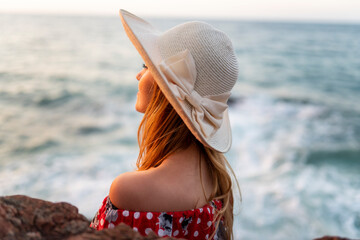 A woman wearing a straw hat is sitting on a rock near the ocean.