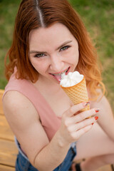 Young beautiful red-haired woman smiling with braces and eating an ice cream cone outdoors. Vertical photo.