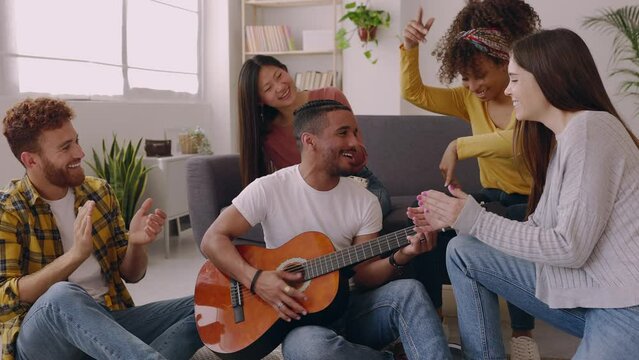 Young group of diverse friends enjoying home party having fun playing the guitar relaxed in the living room. Friendship and leisure concept.