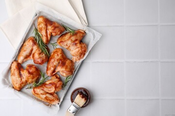 Raw chicken wings, rosemary, marinade and basting brush on light tiled table, flat lay. Space for...