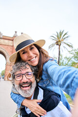 Vertical. Senior gray hair man is giving piggyback ride to mature woman wearing sun hat in happy...