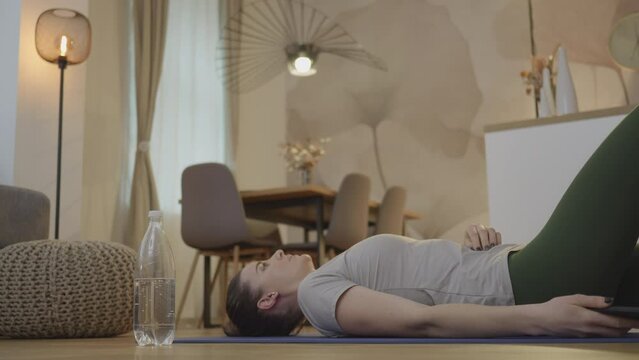 Woman lying on back on yoga mat while exercising and using smartphone. Caucasian girl breathing deeply after workout and checking app on mobile phone side view