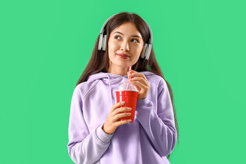 Beautiful young Asian woman in stylish hoodie and headphones holding cup of drink on green background