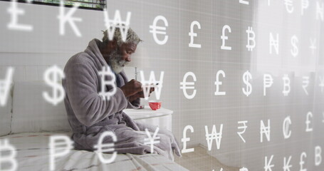 Image of currency symbols, senior african american man sitting on bed and using cellphone - Powered by Adobe