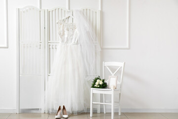 Wedding dress and veil hanging on white folding screen and bridal bouquet on chair in room
