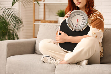 Young woman with weight scales sitting on sofa at home. Weight gain concept