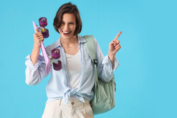 Happy female student with backpack and skateboard pointing at something on blue background