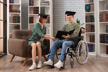 Male graduate in wheelchair with book and his classmate at library