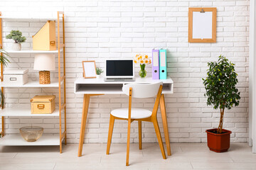 Shelving unit and workplace with bouquet of narcissus flowers and laptop near white brick wall in...