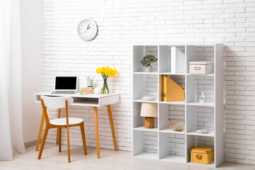 Shelving unit and workplace with bouquet of narcissus flowers and laptop near white brick wall in...