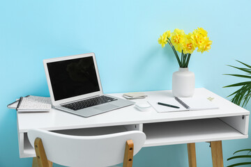 Chair and workplace with bouquet of narcissus flowers and laptop near blue wall in stylish office