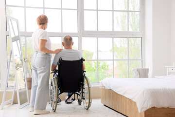 Mature woman with her sick husband in wheelchair at home, back view