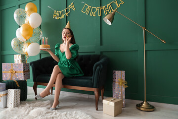 Young woman with Birthday cake and party blower sitting on sofa near green wall