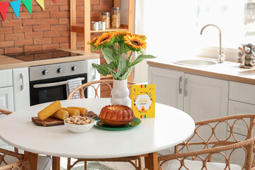 Greeting card with text FESTA JUNINA, traditional food and sunflowers on table in kitchen