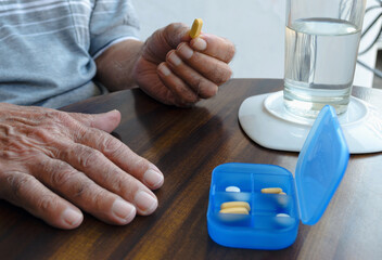 Manos de abuelo sosteniendo una píldora amarilla con vaso de agua y pastillero sobre mesa de...