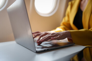 Young Asian business woman in yellow suit uses laptop sitting near window on airplane to do online...