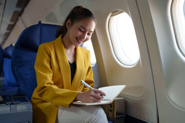 Beautiful successful Asian businesswoman sitting in a plane and working on a notebook. Notes, ideas, business presentation guidelines in first class. Business tourism concept.