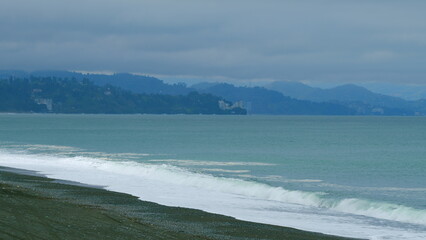 Waves Crashing Against Sea Shore With Mountain Backdrop. Rainy Weather On Sea Coast With Stone...