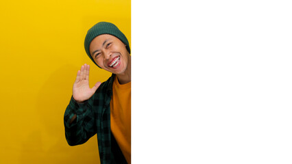 An amazed young Asian man is hiding behind a white wall with an empty space, peeking out and...