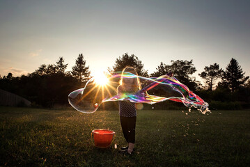 Little girl blowing giant colorful bubble in grassy field at sunset