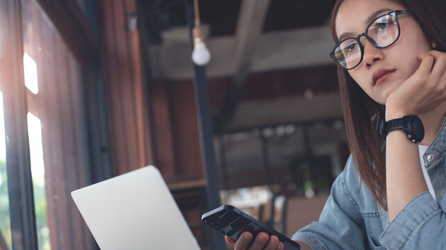 Thoughtful asian young woman looking out of window and thinking on new studying project and working plan, using mobile phone and working on laptop computer, sufing the internet at coffee shop - Powered by Adobe