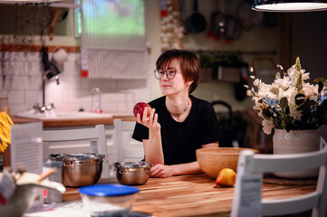 This candid photo captures a young adult in a cozy kitchen, holding an apple and lost in thought. The natural light illuminates their face and glasses, enhancing the thoughtful mood.