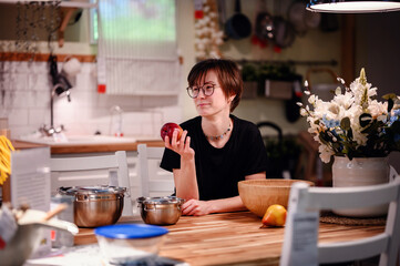 This candid photo captures a young adult in a cozy kitchen, holding an apple and lost in thought....