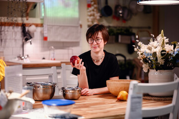 Fototapeta na wymiar This candid photo captures a young adult in a cozy kitchen, holding an apple and lost in thought. The natural light illuminates their face and glasses, enhancing the thoughtful mood.