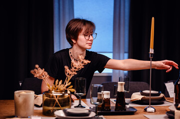 Young adult at an elegantly set dinner table, reaching out to adjust a placement. The sophisticated table setting includes modern tableware and decorative candles, creating a refined dining experience