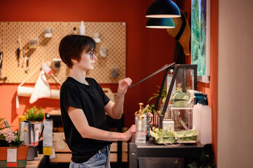 A young adult woman with short hair and glasses is pictured focusing intently on adjusting a plant inside a terrarium.