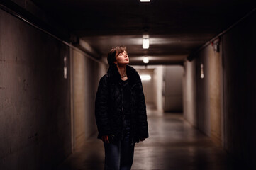 A young woman stands alone in a dimly lit underpass, looking upward with a reflective expression....