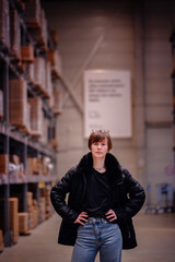 Young woman standing confidently with her hands on her hips in a warehouse environment. Wearing a black puffer jacket and jeans, her determined expression 