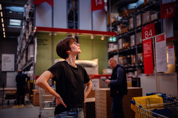 Young woman, hands on hips, looking up thoughtfully in a large retail store. Her casual attire...