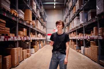 Young woman confidently walking down a warehouse aisle, smartphone in hand, possibly checking...