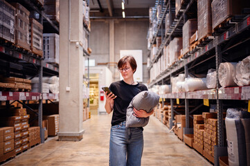 Young woman using her smartphone while shopping in a warehouse, holding a cushion in one arm. Her...
