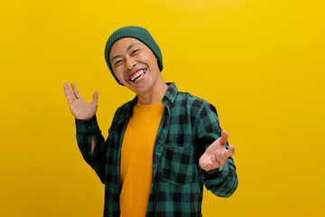 Fototapeta na wymiar Friendly young Asian man, dressed in a beanie hat and casual shirt, pulls his hands towards the camera, making a welcoming gesture, inviting someone inside while standing against a yellow background