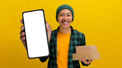 Excited young Asian man, dressed in a beanie hat and casual clothes, showcases a smartphone with an...
