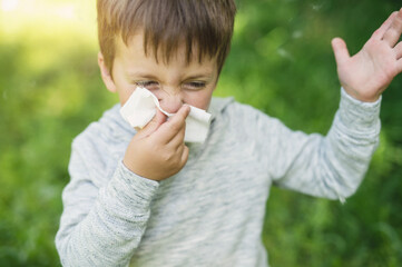 A little boy 4 years old blows his nose into a paper napkin against a background of greenery. Spring exacerbation of allergy to poplar fluff. Allergies in children hay fever