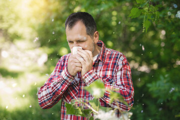 A white European man sneezes and blows his nose into a paper napkin against a green background. Spring exacerbation of allergy to poplar fluff. Allergy in adults hay fever