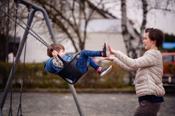 Obraz premium A heartwarming moment of a mother pushing her young son on a swing at a local park. The boy, laughing with glee, reaches out his feet to meet his mother's hands in a playful game. 