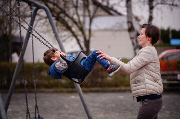 Obraz premium A heartwarming moment of a mother pushing her young son on a swing at a local park. The boy, laughing with glee, reaches out his feet to meet his mother's hands in a playful game. 