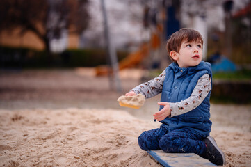 A young boy sits in a sandbox, deeply engaged in play. He holds a sandbox toy, lost in his creative...