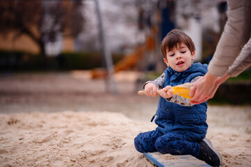 A young boy sits in a sandbox, deeply engaged in play. He holds a sandbox toy, lost in his creative...