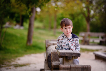 This image beautifully captures a pensive young boy sitting in a wooden structure at a park, surrounded by lush greenery. His contemplative gaze and the natural, 