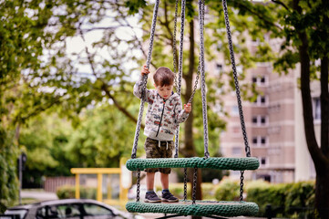 A young boy, attentively focused, stands on a circular swing in an urban park setting. Clad in a patterned hoodie and camo pants, he holds onto the chains, 