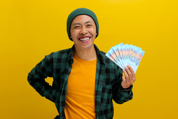 Excited Asian man in a beanie and casual clothes holds banknotes in his hand. Isolated on a bright...