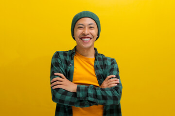 Confident young Asian man, clad in a beanie hat and casual outfit, stands with folded arms, wearing a confident smile as he gazes at the camera while standing against a yellow background