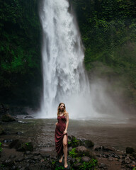 A young blonde woman in a brown long dress at Nungnung waterfall in the tropical forest, Bali, Indonesia