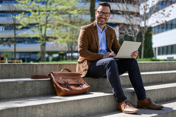 Portrait of smiling manager working over laptop and sitting with bag on steps in city on sunny day