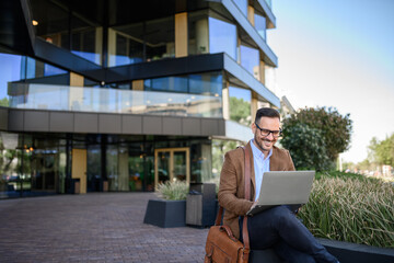 Happy male entrepreneur in glasses trading online over laptop while sitting outside modern building